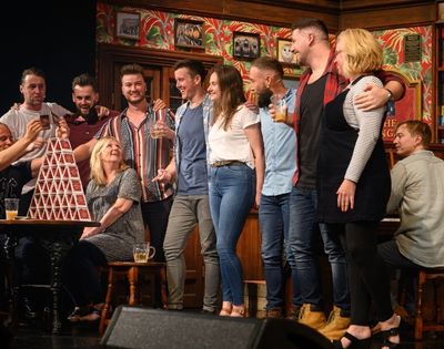 A group of people stand in front of a decorated wall and a piano, with a tall, precariously stacked tower of playing cards on a table in the foreground.