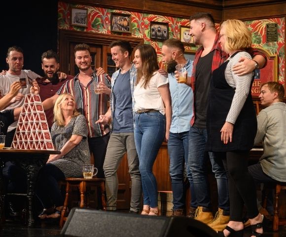 A group of people stand in front of a decorated wall and a piano, with a tall, precariously stacked tower of playing cards on a table in the foreground.