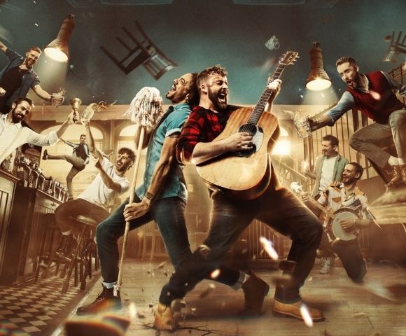 A group of men in a bar, with one playing an acoustic guitar and another singing while holding a mop, while others raise glasses and one plays a banjo.