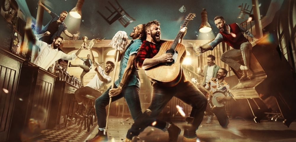 A group of men in a bar, with one playing an acoustic guitar and another singing while holding a mop, while others raise glasses and one plays a banjo.
