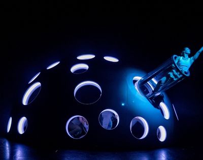 A performer in a cylindrical tube emerges from a large, dark, domed structure with numerous illuminated circular openings, all under intense blue lighting.
