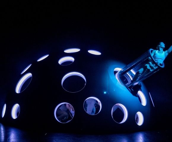 A performer in a cylindrical tube emerges from a large, dark, domed structure with numerous illuminated circular openings, all under intense blue lighting.