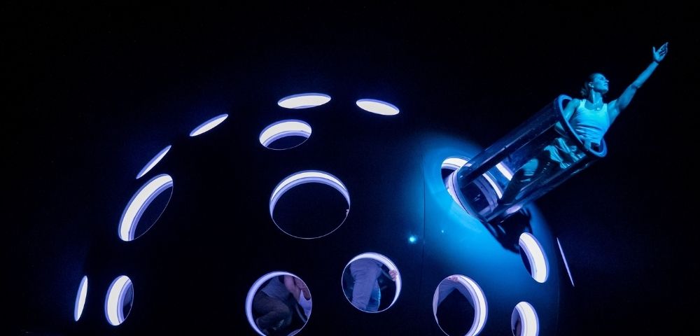 A performer in a cylindrical tube emerges from a large, dark, domed structure with numerous illuminated circular openings, all under intense blue lighting.