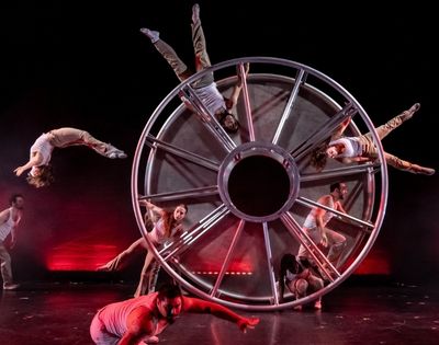 A troupe of acrobats performs with a large, multi-spoked metal wheel on a dark stage with red stage lighting.