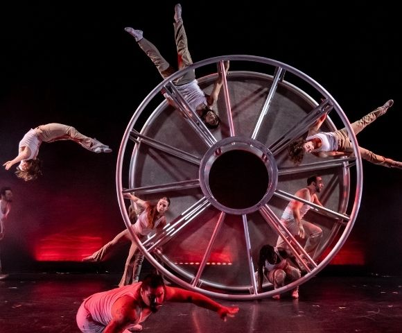 A troupe of acrobats performs with a large, multi-spoked metal wheel on a dark stage with red stage lighting.