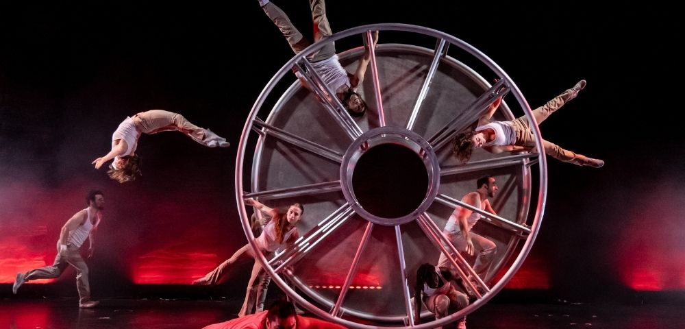 A troupe of acrobats performs with a large, multi-spoked metal wheel on a dark stage with red stage lighting.