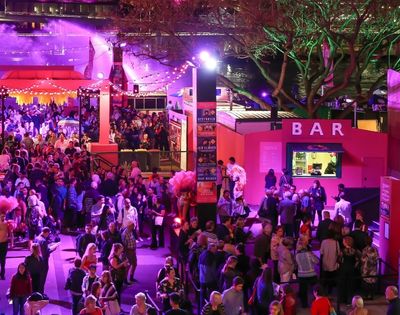 A lively outdoor night market or festival scene with a large crowd of people gathered near a brightly lit pink bar.