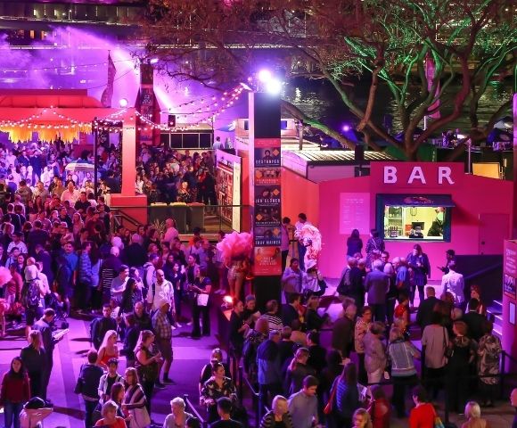A lively outdoor night market or festival scene with a large crowd of people gathered near a brightly lit pink bar.