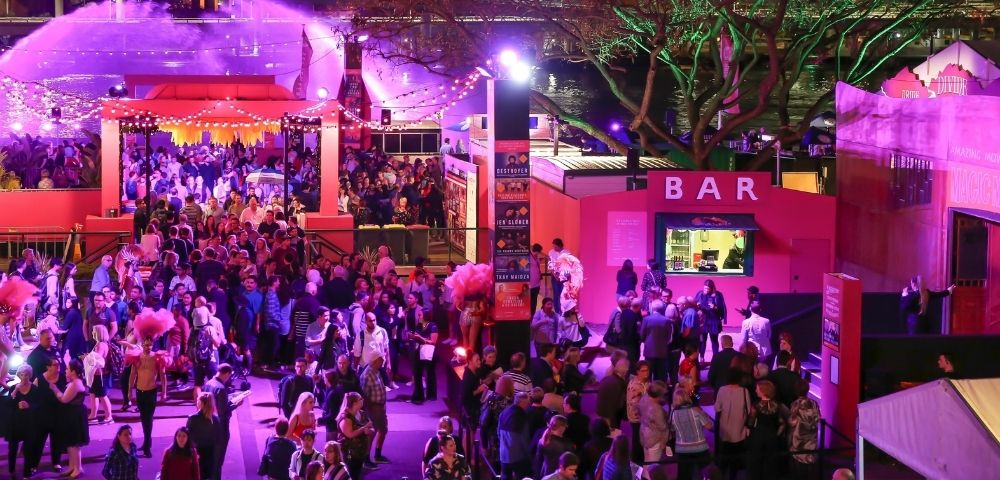 A lively outdoor night market or festival scene with a large crowd of people gathered near a brightly lit pink bar.