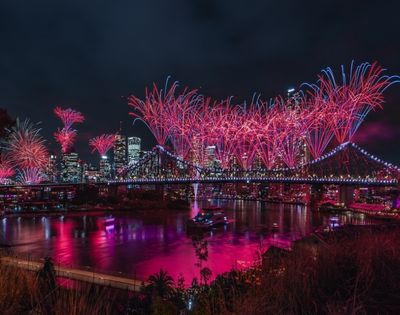Night view of a city skyline with a illuminated bridge covered in red and blue fireworks reflecting in the water below.