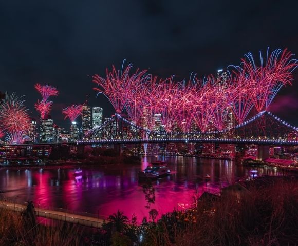 Night view of a city skyline with a illuminated bridge covered in red and blue fireworks reflecting in the water below.