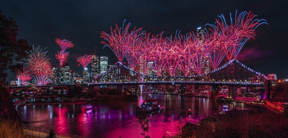 Night view of a city skyline with a illuminated bridge covered in red and blue fireworks reflecting in the water below.