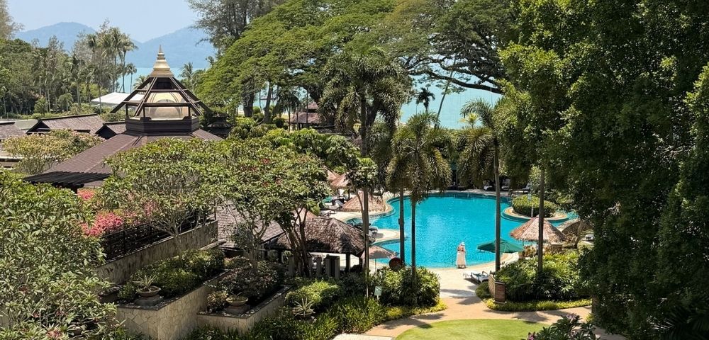 A bright blue swimming pool is surrounded by lush greenery, palm trees, and thatched-roof cabanas, with a multi-level resort building in the background under a clear sky.
