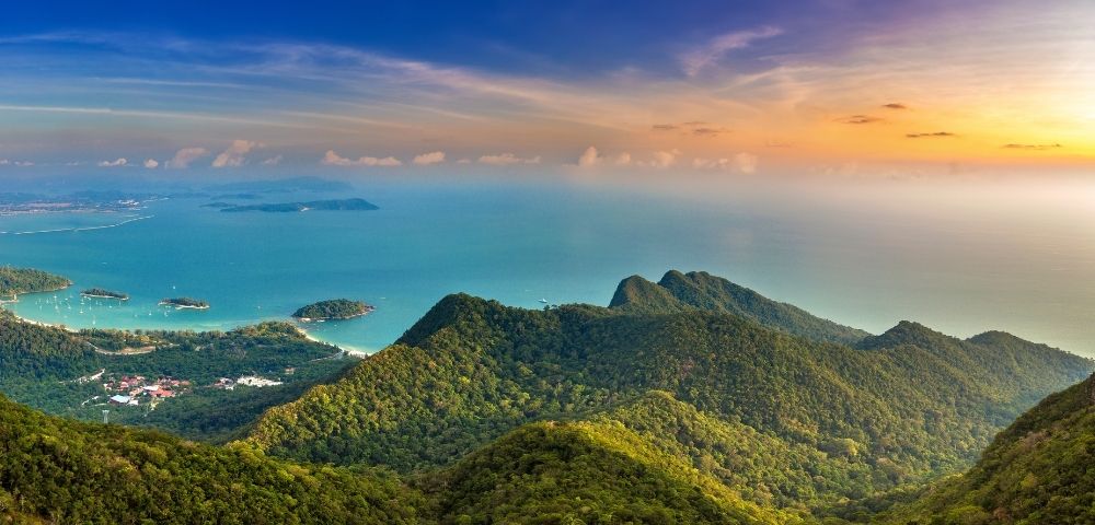 Panoramic aerial view of lush green mountains overlooking a turquoise bay dotted with small islands and sailboats, under a vibrant sunset sky.