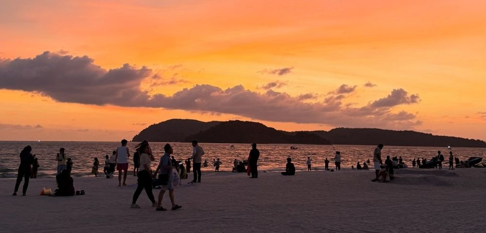 Silhouettes of people enjoying a beach at sunset, with a vibrant orange and yellow sky over dark, hilly islands in the background.