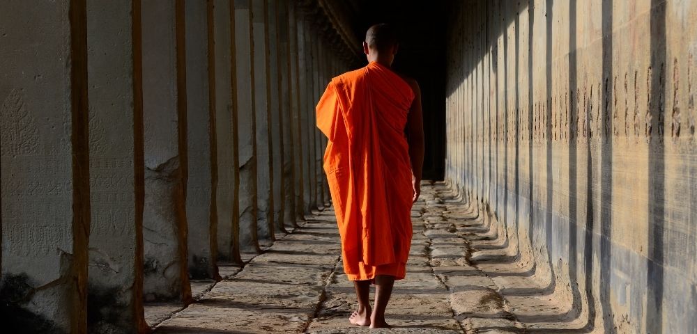 A Buddhist monk in an orange robe walks away from the camera down a long, stone corridor lined with pillars.