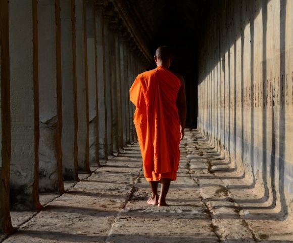 A Buddhist monk in an orange robe walks away from the camera down a long, stone corridor lined with pillars.