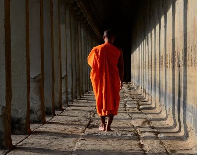A Buddhist monk in an orange robe walks away from the camera down a long, stone corridor lined with pillars.
