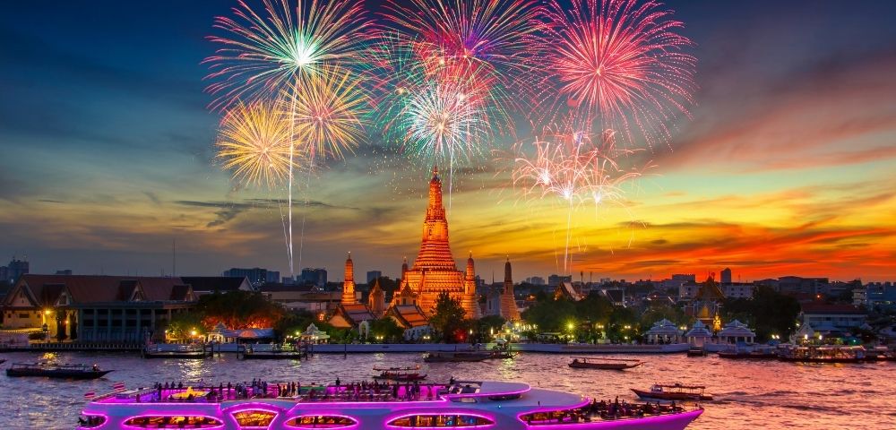 A large, illuminated boat with pink lighting cruises down a river in front of a silhouetted city skyline, featuring a prominent temple with spires. Above the cityscape, a vibrant fireworks display bursts in the twilight sky.