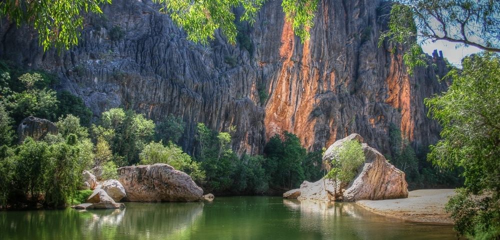 A tranquil, green body of water reflects a dramatic, jagged cliff face. Lush green trees and large boulders line the water's edge. Sunlight illuminates parts of the rocky cliffs with warm orange and brown hues, contrasting with the shadowed grey rock.