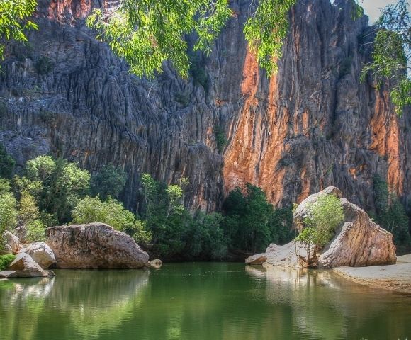 A tranquil, green body of water reflects a dramatic, jagged cliff face. Lush green trees and large boulders line the water's edge. Sunlight illuminates parts of the rocky cliffs with warm orange and brown hues, contrasting with the shadowed grey rock.