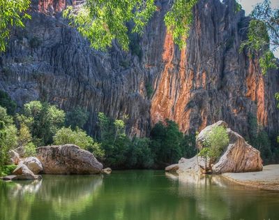 A tranquil, green body of water reflects a dramatic, jagged cliff face. Lush green trees and large boulders line the water's edge. Sunlight illuminates parts of the rocky cliffs with warm orange and brown hues, contrasting with the shadowed grey rock.