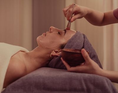 A woman receiving a facial treatment, lying down with a towel on her head while another person applies a mask to her face.