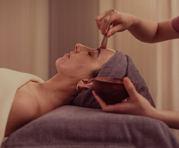A woman receiving a facial treatment, lying down with a towel on her head while another person applies a mask to her face.