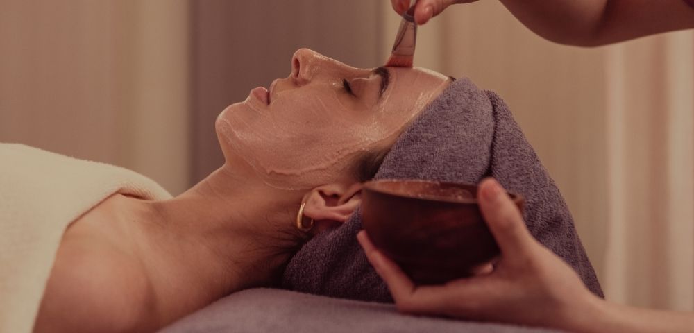 A woman receiving a facial treatment, lying down with a towel on her head while another person applies a mask to her face.