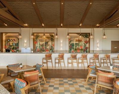 A modern restaurant interior with a bar, featuring high chairs, tables with patterned cushions, and decorative lighting. The bar area is well-stocked and surrounded by tropical-themed decor.