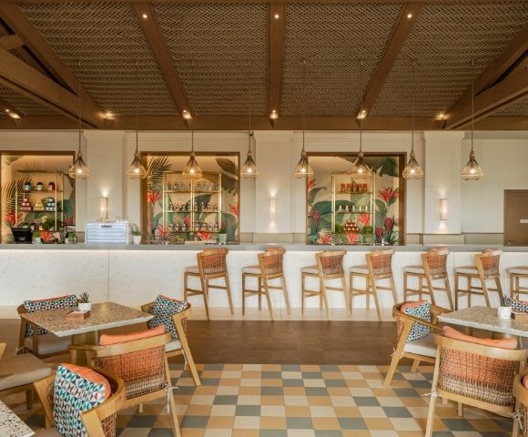 A modern restaurant interior with a bar, featuring high chairs, tables with patterned cushions, and decorative lighting. The bar area is well-stocked and surrounded by tropical-themed decor.