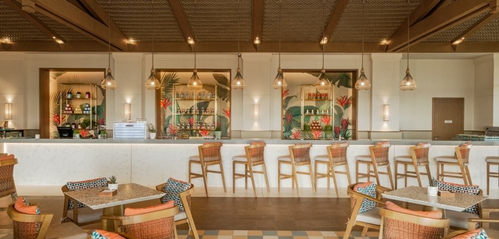 A modern restaurant interior with a bar, featuring high chairs, tables with patterned cushions, and decorative lighting. The bar area is well-stocked and surrounded by tropical-themed decor.