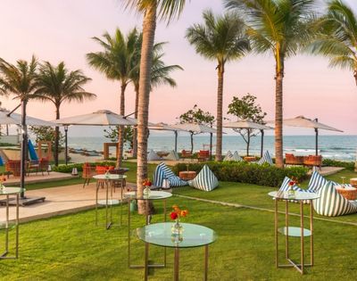 A serene beachside setting featuring palm trees, umbrellas, and modern seating arrangements on a lush lawn. The ocean is visible in the background with a colorful sky at sunset.