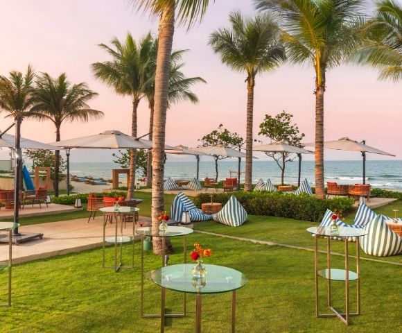 A serene beachside setting featuring palm trees, umbrellas, and modern seating arrangements on a lush lawn. The ocean is visible in the background with a colorful sky at sunset.