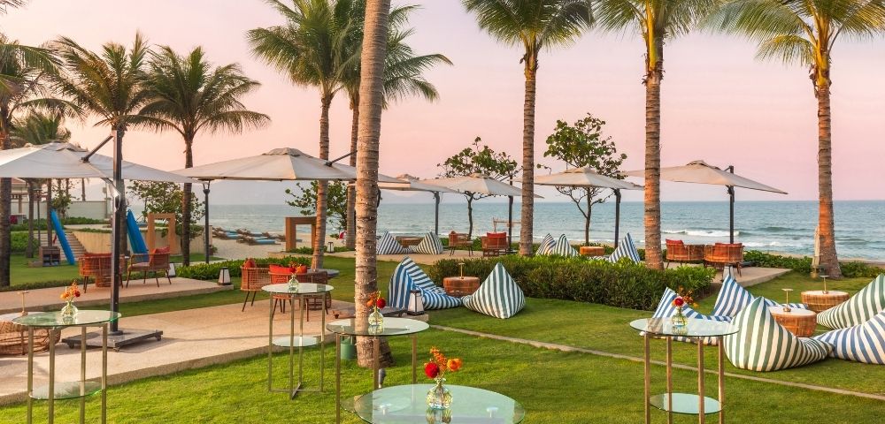 A serene beachside setting featuring palm trees, umbrellas, and modern seating arrangements on a lush lawn. The ocean is visible in the background with a colorful sky at sunset.