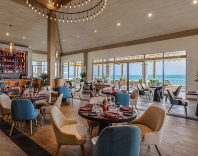 Interior of a restaurant with a view of the ocean, featuring round tables with colorful chairs, a bar area, and large windows allowing natural light in.