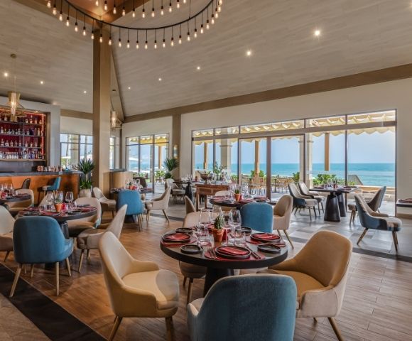 Interior of a restaurant with a view of the ocean, featuring round tables with colorful chairs, a bar area, and large windows allowing natural light in.