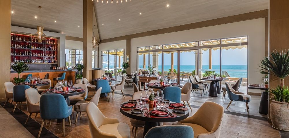 Interior of a restaurant with a view of the ocean, featuring round tables with colorful chairs, a bar area, and large windows allowing natural light in.