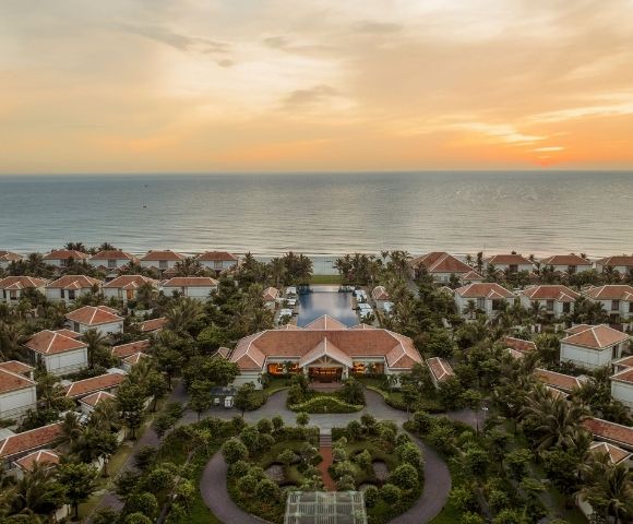 Aerial view of a coastal resort at sunset, featuring numerous white villas surrounded by palm trees and landscaped gardens, with the ocean in the background.