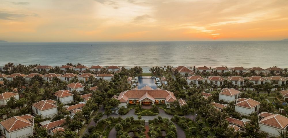 Aerial view of a coastal resort at sunset, featuring numerous white villas surrounded by palm trees and landscaped gardens, with the ocean in the background.