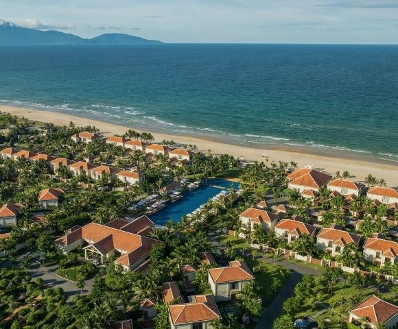 Aerial view of a coastal resort with multiple villas surrounded by greenery and a beach in the background.