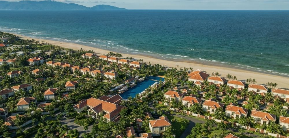 Aerial view of a coastal resort with multiple villas surrounded by greenery and a beach in the background.