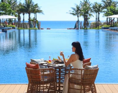 A woman sitting at a table by a pool, enjoying drinks and food with an ocean view.