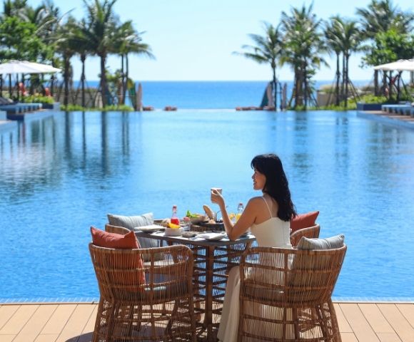 A woman sitting at a table by a pool, enjoying drinks and food with an ocean view.