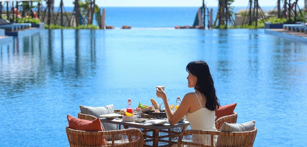 A woman sitting at a table by a pool, enjoying drinks and food with an ocean view.