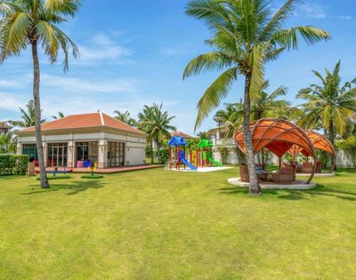 A resort playground with a colorful climbing structure, slides, and seesaws surrounded by lush green grass and tall palm trees, with a modern building and outdoor seating areas in the background under a clear blue sky.