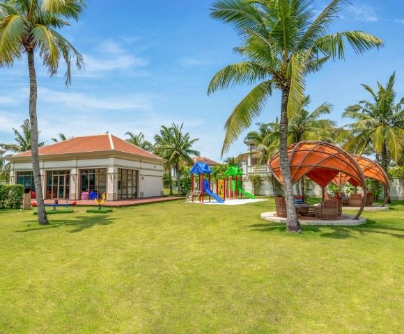 A resort playground with a colorful climbing structure, slides, and seesaws surrounded by lush green grass and tall palm trees, with a modern building and outdoor seating areas in the background under a clear blue sky.