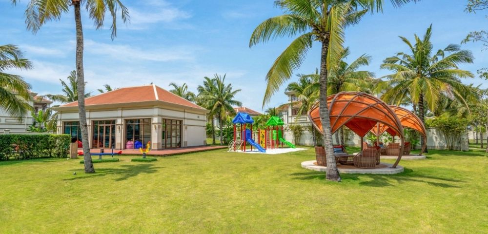 A resort playground with a colorful climbing structure, slides, and seesaws surrounded by lush green grass and tall palm trees, with a modern building and outdoor seating areas in the background under a clear blue sky.