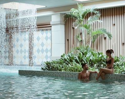 A couple relaxes in a tranquil indoor pool area with a cascading waterfall feature and lush greenery, set against a backdrop of patterned tiles and vertical wooden slats.