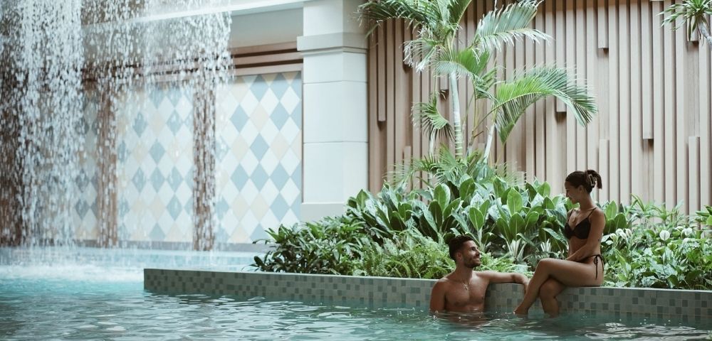 A couple relaxes in a tranquil indoor pool area with a cascading waterfall feature and lush greenery, set against a backdrop of patterned tiles and vertical wooden slats.
